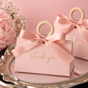 Könnte beinhalten: Zwei hellrosa Geschenkboxen mit einem dezenten geprägten Blumenmuster und dem Text "thank you" in goldener Schrift. Jede Box ist mit einem glatten Holzringgriff und einem passenden hellrosa Satinband versehen, das zu einer Schleife gebunden ist.