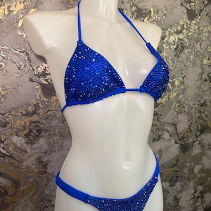 May include: A vibrant royal blue bikini set featuring a triangle top and matching bottoms. Both pieces are densely embellished with sparkling blue crystals that cover the fabric, creating a reflective surface across the entire swimwear set.