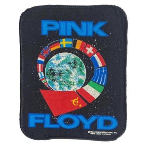 Könnte beinhalten: Ein rechteckiger schwarzer Stoffaufnäher mit dem blauen Schriftzug PINK FLOYD. Das Design zeigt einen zentralen Globus, der von einer kreisförmigen Anordnung verschiedener Nationalflaggen umgeben ist, mit einer roten Flagge mit gelbem Symbol unten.