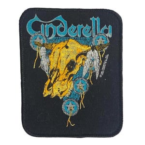 Puede incluir: Un parche rectangular negro bordado que presenta la palabra Cinderella en letra cursiva azul claro en la parte superior. Debajo del texto hay un gráfico de cráneo de buey amarillo adornado con plumas blancas y tres emblemas de estrellas circulares azul claro.
