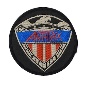 Peut inclure: Un écusson brodé circulaire présentant un motif de bouclier avec une bordure noire. Le bouclier affiche une bande horizontale bleue avec le texte rouge ANTHRAX, au-dessus de rayures verticales rouges et blanches. Un aigle est au sommet.