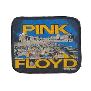 Könnte beinhalten: Ein rechteckiger Stoffaufnäher mit schwarzem Rand, der eine Grafik eines großen Gebäudes hinter einem Feld von Krankenhausbetten zeigt. Der Text PINK FLOYD ist in fetten gelben Buchstaben gedruckt. Das Bild enthält den Text 1988 Promo Productions Inc.