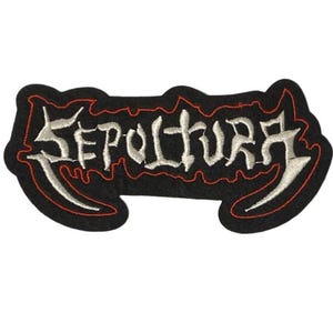Op de afbeelding: Een geborduurde stoffen patch met het woord SEPULTURA in een gestileerd, grillig wit lettertype. De tekst staat op een zwarte achtergrond met een dunne rode gestikte rand die de onregelmatige, organische vorm van de letters volgt.