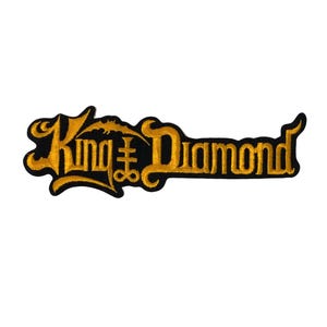 Pode incluir: Um emblema bordado com o texto King Diamond numa fonte dourada estilizada com uma borda preta. O design inclui um símbolo de cruz integrado nas letras, tudo sobre um fundo branco liso.