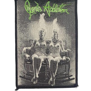 Op de afbeelding: Een rechthoekige geweven patch met een zwart-wit afbeelding van twee figuren die in schommelstoelen zitten, met felgroene geborduurde tekst bovenaan die Jane's Addiction leest. De patch heeft een effen zwarte rand.