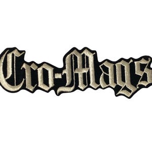 Może przedstawiać: Haftowana naszywka z napisem Cro-Mags wykonanym białą czcionką w stylu gotyckim z czarnym obrysem. Litery są wyszyte gęstym ściegiem, co tworzy wypukłą fakturę na czarnym, materiałowym tle naszywki.