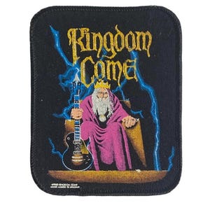 May include: A rectangular embroidered patch with a black background featuring the text Kingdom Come in gold. The design shows a bearded figure in a purple robe and gold crown sitting on a throne, holding a black electric guitar amidst blue lightning.