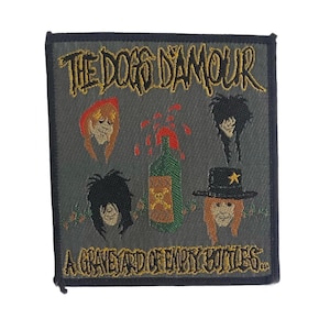 May include: A rectangular embroidered patch with a dark grey background. It features the text THE DOGS D'AMOUR at the top and A GRAVEYARD OF EMPTY BOTTLES at the bottom in yellow. The design includes four illustrated faces and a green bottle.