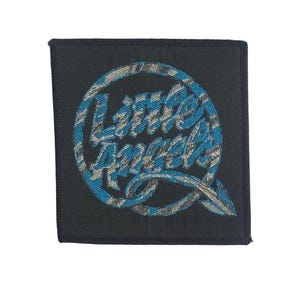 Peut inclure: Un écusson carré en tissu noir avec un motif circulaire brodé en fil bleu et argent. Le texte Little Angels est écrit dans une police cursive stylisée à l'intérieur du cercle, qui se termine par une forme de flèche pointue en bas.