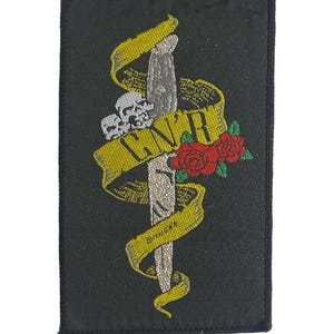 Puede incluir: Un parche rectangular negro bordado que presenta una daga plateada centrada verticalmente. Una cinta amarilla envuelve la daga con el texto GN'R en negro. Una calavera blanca y dos rosas rojas con hojas verdes se encuentran junto a la cinta.