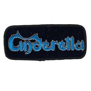 Può includere: Una toppa rettangolare ricamata con uno sfondo in tessuto nero e un bordo nero spesso. La parola Cinderella è cucita con filo blu brillante e un contorno metallico argentato, creando un netto contrasto con la base scura della toppa.