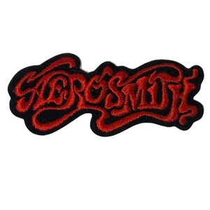 Puede incluir: Un parche bordado que presenta la palabra Aerosmith en una fuente roja estilizada y fluida. Las letras están cosidas con hilo rojo grueso y delineadas sobre un fondo de tela negra sólida, creando un diseño audaz de alto contraste.