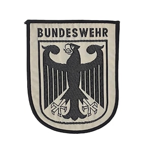 Könnte beinhalten: Ein rechteckiger Stoffaufnäher mit abgerundeten unteren Ecken, der einen schwarzen, gestickten Adler auf cremefarbenem Hintergrund zeigt. Das Wort BUNDESWEHR ist in schwarzen Großbuchstaben oben aufgestickt. Der Aufnäher hat einen schwarzen Rand.