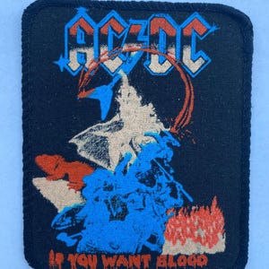 Könnte beinhalten: Ein rechteckiger schwarzer Stoffaufnäher mit dem AC/DC-Logo in Blau und Weiß im oberen Bereich. Unter dem Logo befindet sich eine Grafik einer Figur mit Gitarre und Flammen. Der Text IF YOU WANT BLOOD ist in Orange am unteren Rand aufgedruckt.