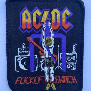 Op de afbeelding: Een rechthoekige zwarte stoffen patch met een geborduurd AC/DC-logo in geel, oranje en rood. Het ontwerp toont een figuur met opgeheven armen, een gitaar, versterkers en de tekst FLICK OF THE SWITCH in oranje letters op een zwarte achtergrond.