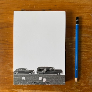 May include: Black and white photograph of a funeral procession with two cars, a hearse and a sedan, driving past a row of headstones.