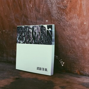 May include: A white notebook with the text "DYING TO DO." printed on the front. The notebook is sitting on a wooden surface. The cover features a black and white image of a skull.