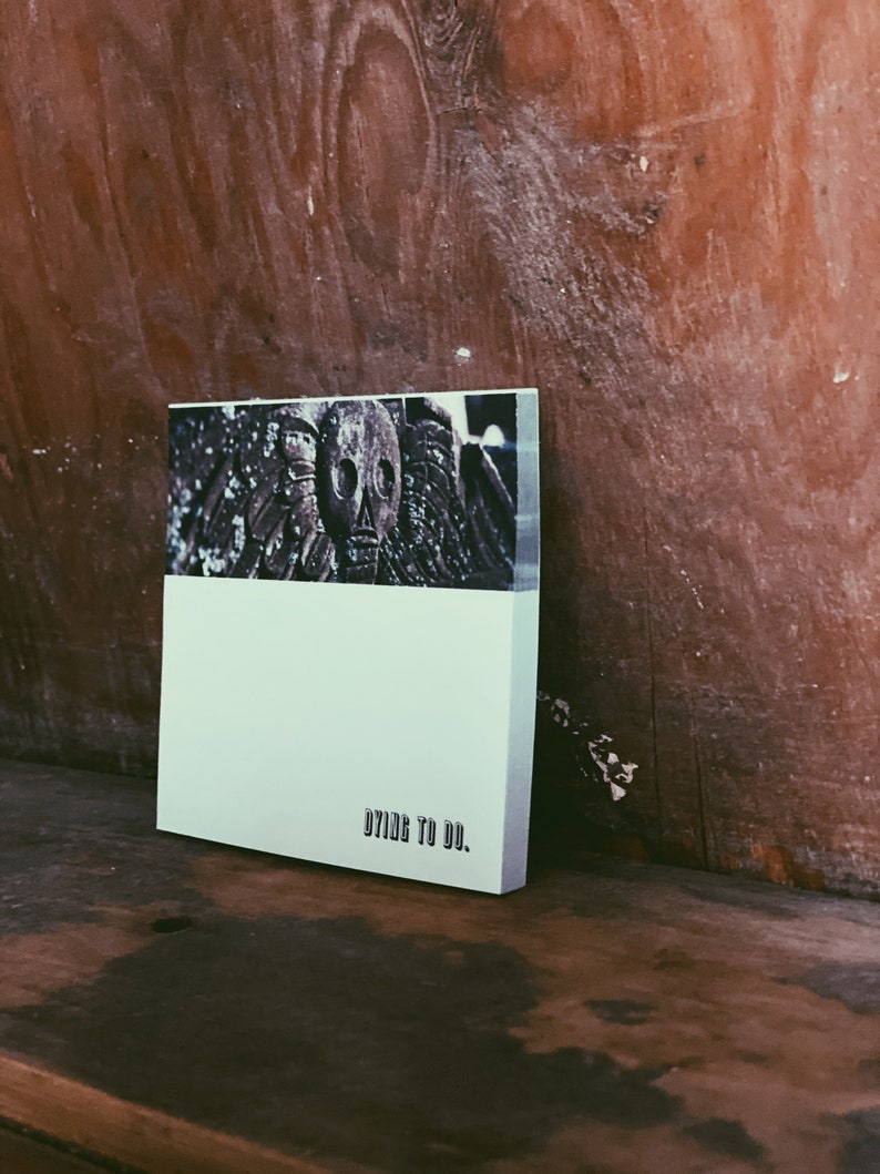 May include: A white notebook with the text "DYING TO DO." printed on the front. The notebook is sitting on a wooden surface. The cover features a black and white image of a skull.
