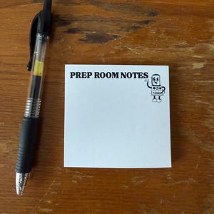 May include: A black and silver pen lies next to a white notepad with the text "PREP ROOM NOTES" and a cartoon character illustration. The pen has a clear barrel with yellow ink visible. The notepad is on a wooden surface.