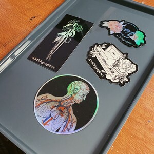 May include: Four holographic stickers with black backgrounds and white outlines. The stickers feature a skull with flowers, a coffin with a rose, a bouquet of flowers, and a human anatomy illustration. The stickers all have the text "AMEKgraphics" on them.
