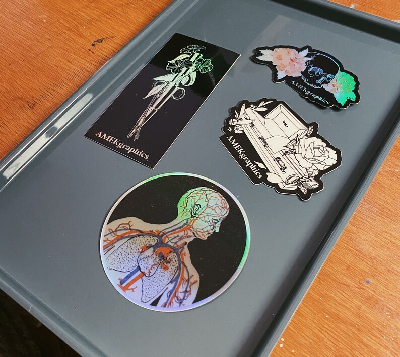 May include: Four holographic stickers with black backgrounds and white outlines. The stickers feature a skull with flowers, a coffin with a rose, a bouquet of flowers, and a human anatomy illustration. The stickers all have the text "AMEKgraphics" on them.