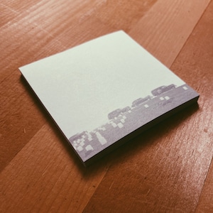 May include: A white sticky note with a faint gray image of a cityscape printed on it. The note is lying on a wooden surface.