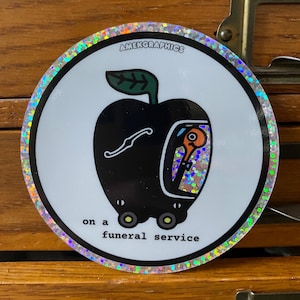 May include: A black and green apple with a glittery orange eye on wheels. The text "on a funeral service" is below the apple. The sticker has a holographic border.