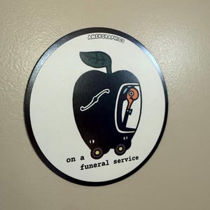 May include: A circular sticker with a black border and a white background. It features a black apple-shaped vehicle with a person inside. The text "AMEKGRAPHICS" is at the top, and "on a funeral service" is at the bottom.