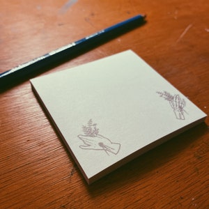 May include: A stack of square, white sticky notes with a purple hand-holding-flowers design in the corners. A blue pencil rests on a wooden surface next to the notepad.