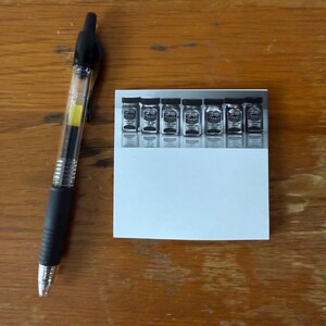 May include: A clear plastic pen with a black grip and a yellow ink cartridge is next to a white card with six small jars printed on it. The jars have black lids and labels. The background is a wooden surface.