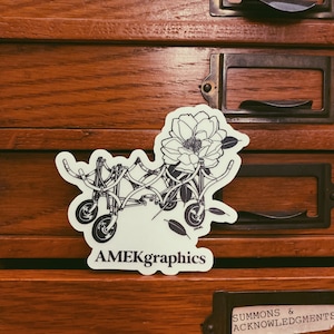 May include: A black and white sticker with a line drawing of a bicycle with a flower on the handlebars. The sticker says "AMEKgraphics" at the bottom.