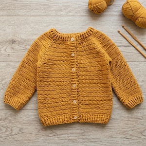 May include: A mustard yellow long-sleeve cardigan with a ribbed texture, featuring a round neckline and a vertical row of six light-colored wooden buttons down the center front. Two balls of matching yarn and two knitting needles are visible.
