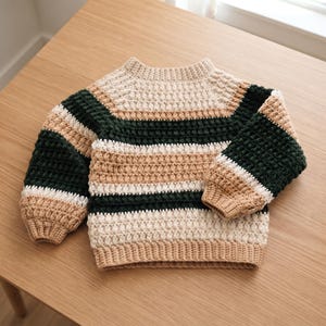 May include: A long-sleeved crochet jumper featuring horizontal stripes in dark green, cream, and tan. The garment is constructed with a textured bobble stitch pattern, a round neckline, and ribbed cuffs and hem in a solid tan yarn colour.