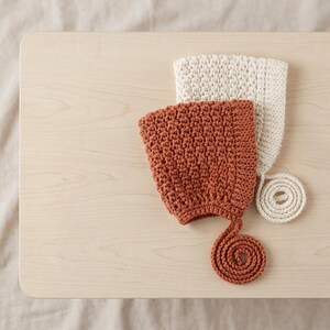 May include: Two handmade crochet bonnets, one in a solid rust orange colour and one in a solid cream colour. Each bonnet features a textured stitch pattern, a structured brim, and a long, coiled crochet tie attached to the bottom edge.