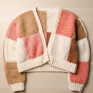 May include: A cropped, long-sleeve cardigan made of crocheted yarn in a patchwork pattern. The garment features alternating square blocks of cream, tan, and coral pink colours, with a ribbed hem and cuffs and an open front design.