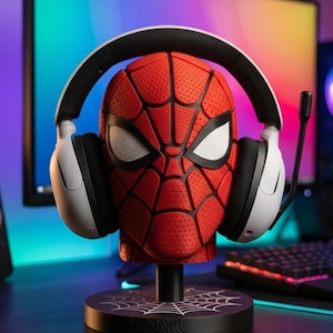 Puede incluir: Un soporte de exhibición con forma de cabeza de Spider-Man en rojo y negro texturizado con lentes oculares blancas. La máscara está montada sobre una base circular negra con un gráfico de telaraña blanca y sostiene auriculares gaming con micrófono.