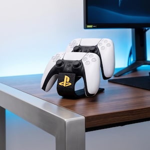 May include: A black plastic controller stand featuring a gold PlayStation logo on the front. The stand holds two white and black wireless game controllers, which are positioned one behind the other in a vertical orientation on the desk surface.