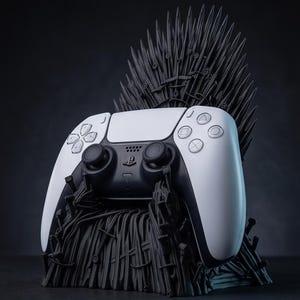 May include: A white and black video game controller resting in a black stand shaped like a throne made of numerous pointed swords. The controller features standard directional buttons, two analog sticks, and a central touchpad on its front surface.