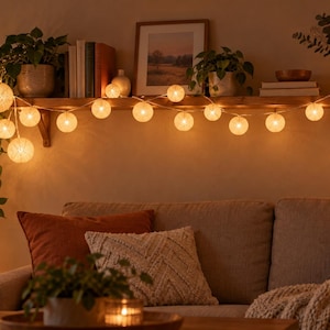 May include: A string of decorative globe lights featuring round, textured, off-white shades that emit a warm, soft glow. The lights are arranged in a horizontal line along a wooden shelf, creating a series of illuminated spheres across the wall.