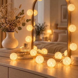May include: A string of warm white spherical cotton ball lights draped around an arched mirror with a gold frame. The illuminated globes feature a textured, woven appearance, casting a soft glow across the surface of the wooden dresser.
