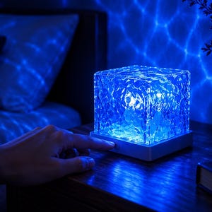 Puede incluir: Una lámpara de forma cuadrada con una pantalla de cristal texturizado y cristalino que emite una luz azul vibrante. La lámpara cuenta con una base blanca con un pequeño botón de encendido circular en la parte frontal, proyectando un patrón ondulado.