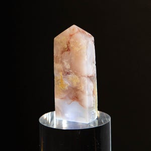 Peut inclure: Une tour en cristal polie avec un sommet pointu, présentant des inclusions translucides blanches, rose pâle et brun orangé terreux à travers la pierre, exposée debout sur un socle cylindrique en acrylique transparent.