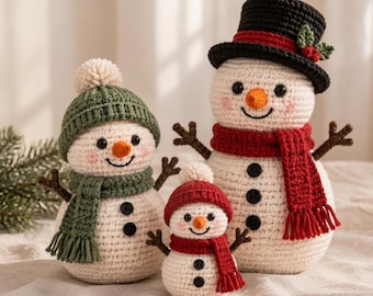 Puede incluir: Tres muñecos de nieve tejidos a ganchillo de diferentes tamaños. El más grande lleva un sombrero de copa negro con borde rojo y acebo, y una bufanda roja. El mediano tiene un gorro verde con pompón y bufanda verde. El más pequeño lleva un gorro rojo.
