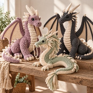 Könnte beinhalten: Drei gehäkelte Drachen-Plüschtiere. Einer ist malvenfarben mit cremefarbenen Akzenten, einer ist dunkel anthrazitgrau mit cremefarbenen Akzenten und einer ist ein langer, schlangenförmiger hellgrüner Drache mit cremefarbenen Details, langen Schnurrhaaren und einem gefransten Schwanz.