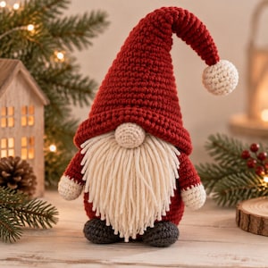 May include: A crochet gnome figurine featuring a tall, pointed red hat with a white pom-pom at the tip. The gnome has a round beige nose, a long cream-coloured yarn beard, red sleeves with white cuffs, and dark grey feet at the base of the body.