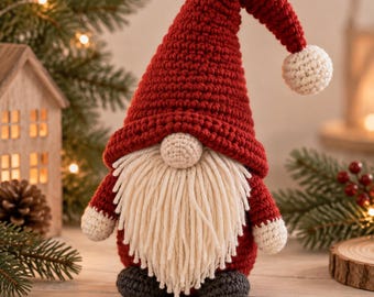 Puede incluir: Una figura de gnomo tejida a ganchillo con un gorro rojo alto y puntiagudo que termina en un pompón blanco. El gnomo tiene una nariz redonda de color beige, una larga barba de hilo crema, mangas rojas con puños blancos y pies gris oscuro.