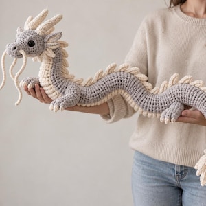 Op de afbeelding: Een lang, gehaakt draak-speeltje met een grijs lichaam en crèmekleurige accenten. Het wezen heeft een gedetailleerde kop met grote zwarte ogen, lange crèmekleurige snorharen en een reeks crèmekleurige stekels langs de gehele rug.