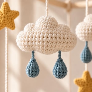 May include: A crochet mobile featuring a central off-white cloud with two dangling blue raindrops suspended by yarn. To the left and right, additional crochet elements include a yellow star and another white cloud with a blue raindrop attached.