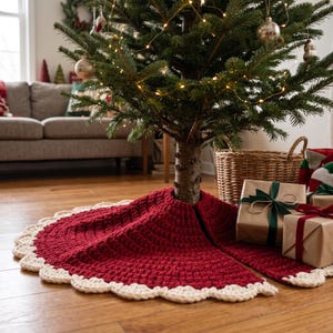 Puede incluir: Una falda circular de ganchillo para &aacute;rbol de Navidad en color rojo oscuro con un borde festoneado grueso de color crema. La falda rodea la base de un pino verde, con una abertura visible y un acabado de hilo grueso y texturizado.