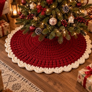 Könnte beinhalten: Ein runder, dunkelroter gehäkelter Weihnachtsbaumteppich mit einem dicken, strukturierten Maschenmuster und einem breiten, cremefarbenen Wellenrand. Der Teppich liegt am Fuß eines mit warmweißen Lichtern geschmückten Tannenbaums.